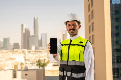 Showing a blank black screen, Saudi national construction projects, interest in urban development by the hands of Saudi engineers, a Saudi Gulf Arab engineer wearing a work jacket and a white protective helmet holding a mobile phone, integrating technology into architecture, following up on the engineering project plan