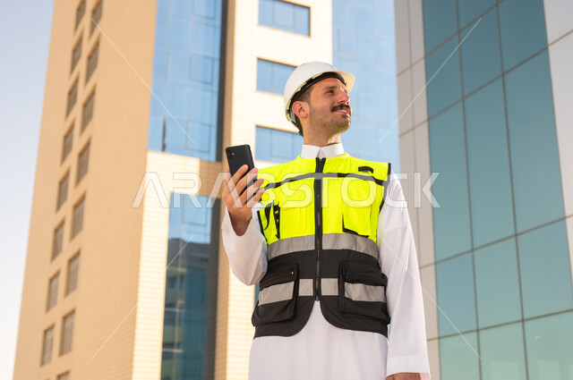 مشاريع إعمارية سعودية وطنية، الاهتمام بالتطور العمراني بسواعد المهندسين السعوديين, مهندس عربي خليجي سعودي يرتدي سترة العمل وخوذة الحماية البيضاء يمسك بيده الهاتف المحمول, دمج التقنية في الهندسة المعمارية, متابعة مخطط المشروع الهندسي