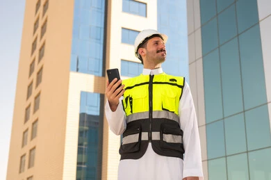 Saudi national construction projects, interest in urban development by Saudi engineers, a Saudi Gulf Arab engineer wearing a work jacket and a white protective helmet holding a mobile phone, integrating technology into architecture, following up on the engineering project plan