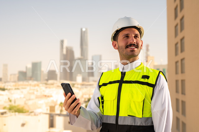 Saudi national construction projects, interest in urban development by Saudi engineers, a Saudi Gulf Arab engineer wearing a work jacket and a white protective helmet holding a mobile phone, integrating technology into architecture, following up on the engineering project plan