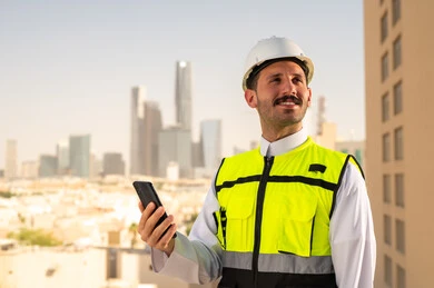 Saudi national construction projects, interest in urban development by Saudi engineers, a Saudi Gulf Arab engineer wearing a work jacket and a white protective helmet holding a mobile phone, integrating technology into architecture, following up on the engineering project plan