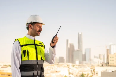 سهولة التواصل مع الزملاء, مشاريع إعمارية سعودية وطنية، الاهتمام بالتطور العمراني بسواعد المهندسين السعوديين, مهندس عربي خليجي سعودي يرتدي سترة العمل وخوذة الحماية البيضاء يمسك بيده جهاز لاسلكي, دمج التقنية في الهندسة المعمارية, متابعة مخطط المشروع الهندسي