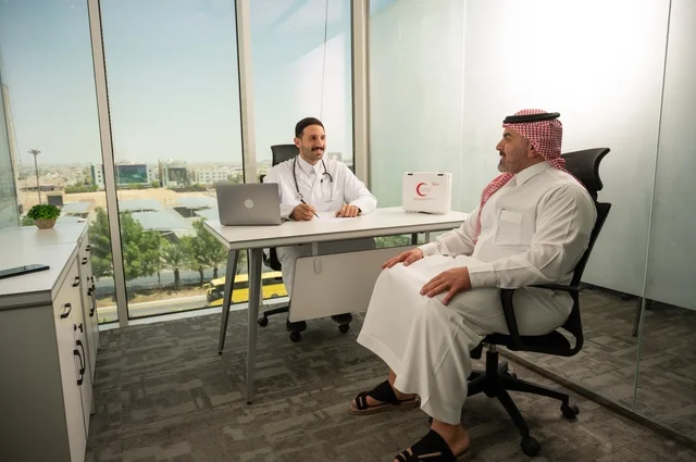 Recording patient cases on the computer, an Arab Gulf Saudi doctor wearing a white coat is explaining the medical condition to a patient wearing a ghutrah and traditional thobe, conversing with patients to uncover the condition, providing consultations in medical centers, following up on the medical records of patients, working in the health sectors.