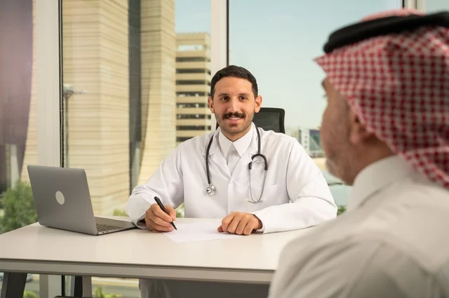 متابعة السجل الطبي للمراجعين, تسجيل حالات المرضى على جهاز الحاسوب, طبيب عربي خليجي سعودي يرتدي المعطف الأبيض يقوم بشرح الحالة المرضية لمريض يرتدي الشماغ والثوب التقليدي، الحوار مع المرضى لكشف الحالة، تقديم الاستشارات في المراكز الطبيه, العمل في القطاعات الصحية