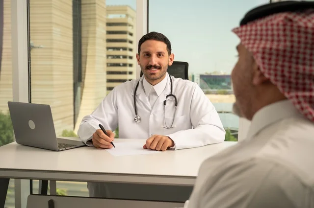 متابعة السجل الطبي للمراجعين, تسجيل حالات المرضى على جهاز الحاسوب, طبيب عربي خليجي سعودي يرتدي المعطف الأبيض يقوم بشرح الحالة المرضية لمريض يرتدي الشماغ والثوب التقليدي، الحوار مع المرضى لكشف الحالة، تقديم الاستشارات في المراكز الطبيه, العمل في القطاعات الصحية