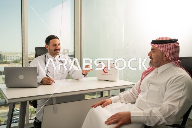 تسجيل حالات المرضى على جهاز الحاسوب, طبيب عربي خليجي سعودي يرتدي المعطف الأبيض يقوم بشرح الحالة المرضية لمريض يرتدي الشماغ والثوب التقليدي، الحوار مع المرضى لكشف الحالة، تقديم الاستشارات في المراكز الطبيه, متابعة السجل الطبي للمراجعين, العمل في القطاعات الصحية