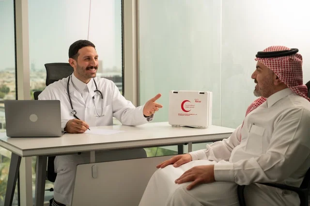 Recording patient cases on the computer, an Arab Gulf Saudi doctor wearing a white coat is explaining the medical condition to a patient wearing a ghutrah and traditional thobe, conversing with patients to uncover the condition, providing consultations in medical centers, following up on the medical records of patients, working in the health sectors.