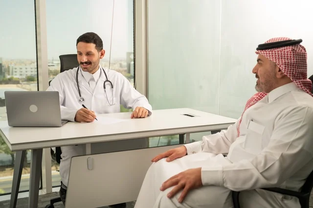 تسجيل حالات المرضى على جهاز الحاسوب, طبيب عربي خليجي سعودي يرتدي المعطف الأبيض يقوم بشرح الحالة المرضية لمريض يرتدي الشماغ والثوب التقليدي، الحوار مع المرضى لكشف الحالة، تقديم الاستشارات في المراكز الطبيه, متابعة السجل الطبي للمراجعين, العمل في القطاعات الصحية