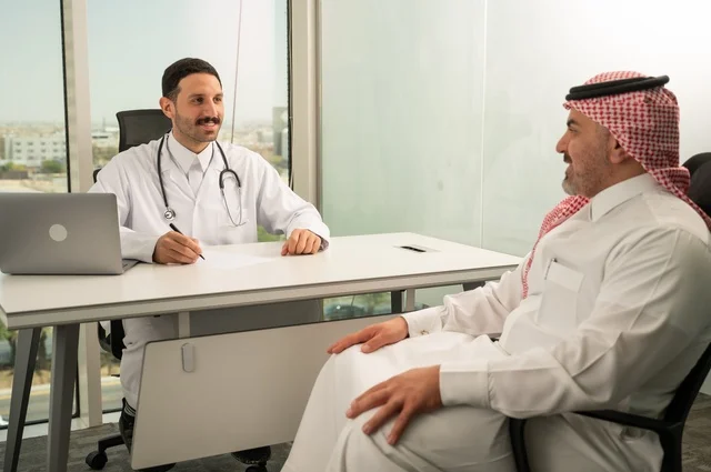 Dialogue with patients to determine their condition, provide consultations in medical centers, follow up on patients' medical records, work in the health sector, record patients' cases on the computer, a Saudi Arabian Gulf doctor wearing a white coat explains the medical condition of a patient wearing a traditional shemagh and thobe