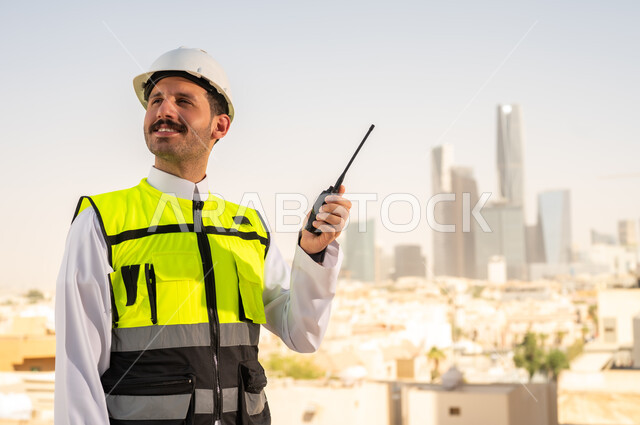 سهولة التواصل مع الزملاء, مشاريع إعمارية سعودية وطنية، الاهتمام بالتطور العمراني بسواعد المهندسين السعوديين, مهندس عربي خليجي سعودي يرتدي سترة العمل وخوذة الحماية البيضاء يمسك بيده جهاز لاسلكي, دمج التقنية في الهندسة المعمارية, متابعة مخطط المشروع الهندسي