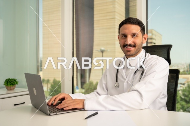 متابعة حالات المرضى عبر الاجهزة و التقنيات الحديثة, استخدام جهاز الحاسوب في الطب في تسجيل الكشوفات والحالات، طبيب عربي خليجي سعودي مبتسم يرتدي المعطف الأبيض يجلس في عيادته وينظر إلى الكاميرا بإيماءات الثقة بالنفس، تسجيل التقارير والحالات الصحيه
