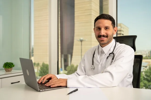 Following up on patients' cases through modern devices and technologies, using computers in medicine to record examinations and cases, a smiling Saudi Arabian Gulf doctor wearing a white coat sitting in his clinic and looking at the camera with gestures of self-confidence, recording reports and health cases
