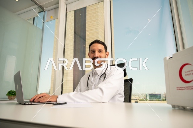 تسجيل التقارير والحالات الصحيه، متابعة حالات المرضى عبر الاجهزة و التقنيات الحديثة, استخدام حقيبة الاسعافات الاولية وجهاز الحاسوب في الطب في تسجيل الحالات، طبيب عربي خليجي سعودي مبتسم يرتدي المعطف الأبيض يجلس في عيادته وينظر إلى الكاميرا بإيماءات الثقة بالنفس
