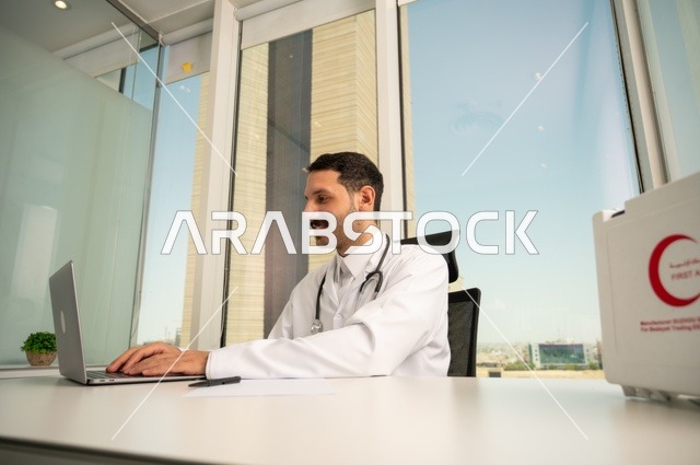 Using a first aid kit and a computer in medicine to record cases, record reports and health conditions, and follow up on patients' cases using modern devices and technologies. A smiling Saudi Arabian Gulf doctor wearing a white coat sits in his clinic.