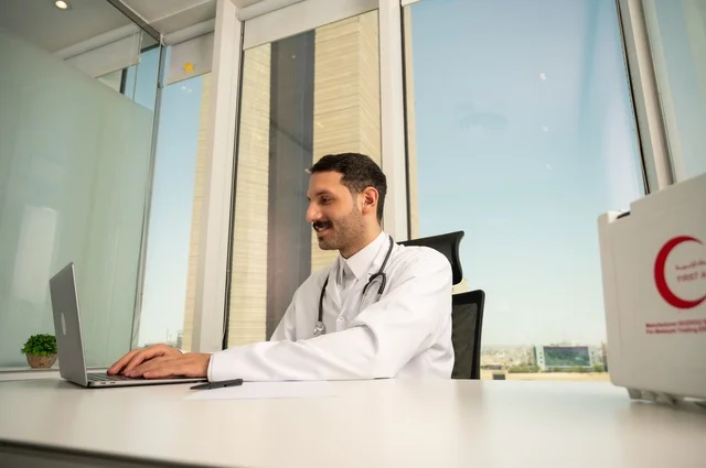 Using a first aid kit and a computer in medicine to record cases, record reports and health conditions, and follow up on patients' cases using modern devices and technologies. A smiling Saudi Arabian Gulf doctor wearing a white coat sits in his clinic.