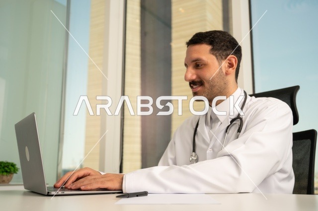 Using a first aid kit and a computer in medicine to record cases, record reports and health conditions, and follow up on patients' cases using modern devices and technologies. A smiling Saudi Arabian Gulf doctor wearing a white coat sits in his clinic.