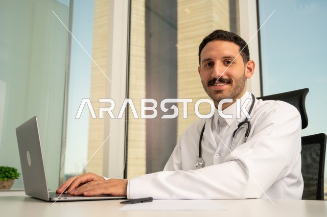متابعة حالات المرضى عبر الاجهزة و التقنيات الحديثة, استخدام جهاز الحاسوب في الطب في تسجيل الكشوفات والحالات، طبيب عربي خليجي سعودي مبتسم يرتدي المعطف الأبيض يجلس في عيادته وينظر إلى الكاميرا بإيماءات الثقة بالنفس، تسجيل التقارير والحالات الصحيه