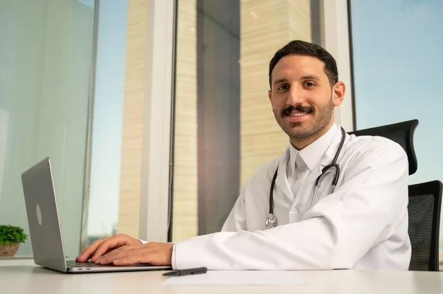 متابعة حالات المرضى عبر الاجهزة و التقنيات الحديثة, استخدام جهاز الحاسوب في الطب في تسجيل الكشوفات والحالات، طبيب عربي خليجي سعودي مبتسم يرتدي المعطف الأبيض يجلس في عيادته وينظر إلى الكاميرا بإيماءات الثقة بالنفس، تسجيل التقارير والحالات الصحيه