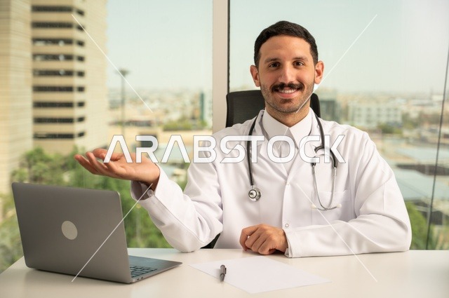 متابعة حالات المرضى عبر الاجهزة و التقنيات الحديثة, استخدام جهاز الحاسوب في الطب في تسجيل الكشوفات والحالات، طبيب عربي خليجي سعودي مبتسم يرتدي المعطف الأبيض يجلس في عيادته وينظر إلى الكاميرا بإيماءات الثقة بالنفس، تسجيل التقارير والحالات الصحيه