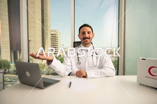 تسجيل التقارير والحالات الصحيه، متابعة حالات المرضى عبر الاجهزة و التقنيات الحديثة, استخدام حقيبة الاسعافات الاولية وجهاز الحاسوب في الطب في تسجيل الحالات، طبيب عربي خليجي سعودي مبتسم يرتدي المعطف الأبيض يجلس في عيادته وينظر إلى الكاميرا بإيماءات الثقة بالنفس