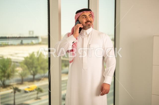 Using a modern and advanced technical device, a Saudi Arabian Gulf man wearing traditional thobe making a phone call via mobile phone, architectural progress and development in the Gulf countries, background of towers and skyscrapers in the city of Riyadh
