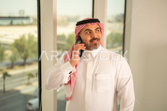 Using a modern and advanced technical device, a Saudi Arabian Gulf man wearing traditional thobe making a phone call via mobile phone, architectural progress and development in the Gulf countries, background of towers and skyscrapers in the city of Riyadh