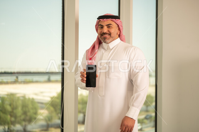 Blank black screen, modern and advanced technical device, Saudi Arabian Gulf man wearing traditional thobe and shemagh using mobile phone, doing remote work, background of towers and skyscrapers in Riyadh