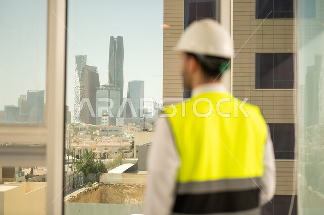 مشاريع إعمارية سعودية وطنية، مفهوم الهندسة المعمارية، متابعة مخطط المشروع الهندسي وتدقيقه, الاهتمام بالتطور العمراني بسواعد المهندسين السعوديين, صورة من الخلف لمهندس عربي خليجي سعودي يرتدي سترة العمل وخوذة الحماية البيضاء يشاهد أبراج مدينة الرياض