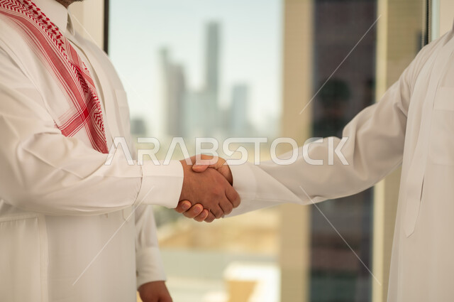 الشراكة في الأعمال والمشاريع الهندسية، صداقات وتعارف وبناء علاقات اجتماعية، عقد صفقات عمل جديدة ناجحة بين رجال الأعمال، صورة مقربة لايدي رجلين سعوديين عربيين خليجيين يرتديان الزي التقليدي يقومان بمصافحة بعضهما، توسيع مجالات العمل