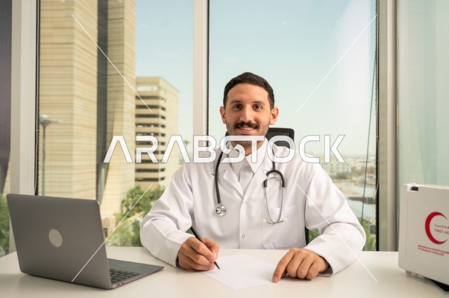 النظر إلى الكاميرا بإيماءات السرور، العمل في القطاعات الصحية، استخدام حقيبة الاسعافات الاولية وجهاز الحاسوب في الطب في تسجيل الحالات، تسجيل التقارير والحالات الصحيه, طبيب عربي خليجي سعودي مبتسم يرتدي المعطف الأبيض يجلس في عيادته