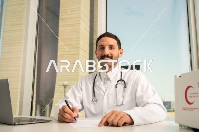 النظر إلى الكاميرا بإيماءات السرور، العمل في القطاعات الصحية، استخدام حقيبة الاسعافات الاولية وجهاز الحاسوب في الطب في تسجيل الحالات، تسجيل التقارير والحالات الصحيه, طبيب عربي خليجي سعودي مبتسم يرتدي المعطف الأبيض يجلس في عيادته