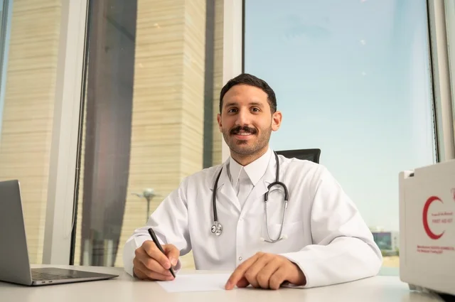 Looking at the camera with gestures of joy, working in the health sectors, using a first aid kit and a computer in medicine to record cases, documenting reports and health cases, a smiling Arab Gulf Saudi doctor wearing a white coat sitting in his clinic.