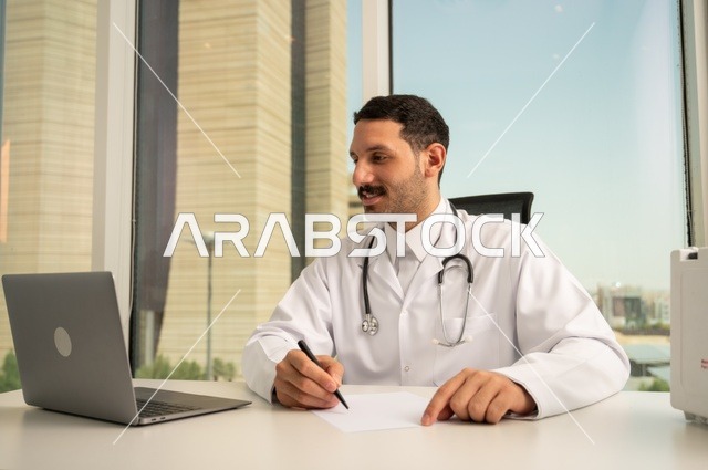 العمل في القطاعات الصحية، استخدام حقيبة الاسعافات الاولية وجهاز الحاسوب في الطب في تسجيل الحالات، تسجيل التقارير والحالات الصحيه، متابعة حالات المرضى عبر الاجهزة و التقنيات الحديثة, طبيب عربي خليجي سعودي مبتسم يرتدي المعطف الأبيض يجلس في عيادته