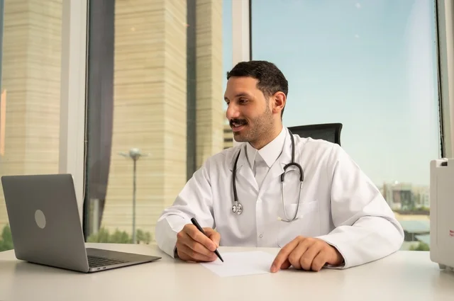 العمل في القطاعات الصحية، استخدام حقيبة الاسعافات الاولية وجهاز الحاسوب في الطب في تسجيل الحالات، تسجيل التقارير والحالات الصحيه، متابعة حالات المرضى عبر الاجهزة و التقنيات الحديثة, طبيب عربي خليجي سعودي مبتسم يرتدي المعطف الأبيض يجلس في عيادته