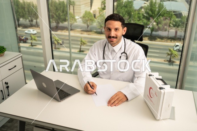 النظر إلى الكاميرا بإيماءات السرور، العمل في القطاعات الصحية، استخدام حقيبة الاسعافات الاولية وجهاز الحاسوب في الطب في تسجيل الحالات، تسجيل التقارير والحالات الصحيه, طبيب عربي خليجي سعودي مبتسم يرتدي المعطف الأبيض يجلس في عيادته