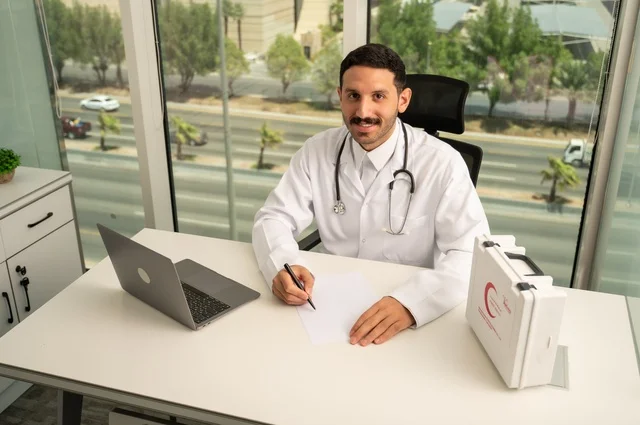النظر إلى الكاميرا بإيماءات السرور، العمل في القطاعات الصحية، استخدام حقيبة الاسعافات الاولية وجهاز الحاسوب في الطب في تسجيل الحالات، تسجيل التقارير والحالات الصحيه, طبيب عربي خليجي سعودي مبتسم يرتدي المعطف الأبيض يجلس في عيادته