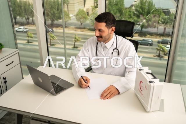 العمل في القطاعات الصحية، استخدام حقيبة الاسعافات الاولية وجهاز الحاسوب في الطب في تسجيل الحالات، تسجيل التقارير والحالات الصحيه، متابعة حالات المرضى عبر الاجهزة و التقنيات الحديثة, طبيب عربي خليجي سعودي مبتسم يرتدي المعطف الأبيض يجلس في عيادته