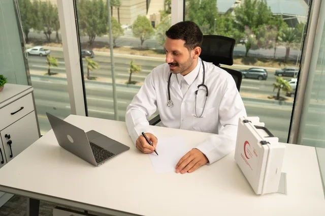 العمل في القطاعات الصحية، استخدام حقيبة الاسعافات الاولية وجهاز الحاسوب في الطب في تسجيل الحالات، تسجيل التقارير والحالات الصحيه، متابعة حالات المرضى عبر الاجهزة و التقنيات الحديثة, طبيب عربي خليجي سعودي مبتسم يرتدي المعطف الأبيض يجلس في عيادته