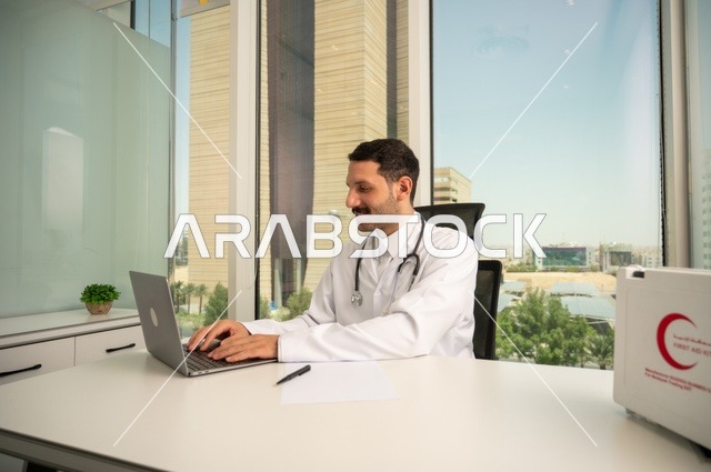 استخدام حقيبة الاسعافات الاولية وجهاز الحاسوب في الطب في تسجيل الحالات، تسجيل التقارير والحالات الصحيه، متابعة حالات المرضى عبر الاجهزة و التقنيات الحديثة, طبيب عربي خليجي سعودي مبتسم يرتدي المعطف الأبيض يجلس في عيادته 