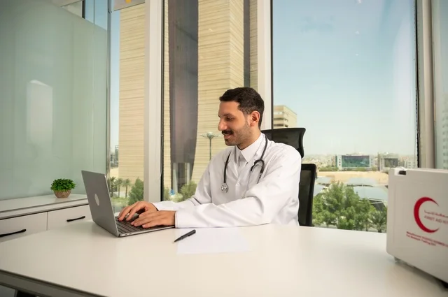 استخدام حقيبة الاسعافات الاولية وجهاز الحاسوب في الطب في تسجيل الحالات، تسجيل التقارير والحالات الصحيه، متابعة حالات المرضى عبر الاجهزة و التقنيات الحديثة, طبيب عربي خليجي سعودي مبتسم يرتدي المعطف الأبيض يجلس في عيادته 