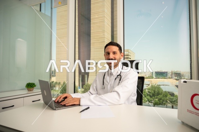 تسجيل التقارير والحالات الصحيه، متابعة حالات المرضى عبر الاجهزة و التقنيات الحديثة, استخدام حقيبة الاسعافات الاولية وجهاز الحاسوب في الطب في تسجيل الحالات، طبيب عربي خليجي سعودي مبتسم يرتدي المعطف الأبيض يجلس في عيادته وينظر إلى الكاميرا بإيماءات الثقة بالنفس