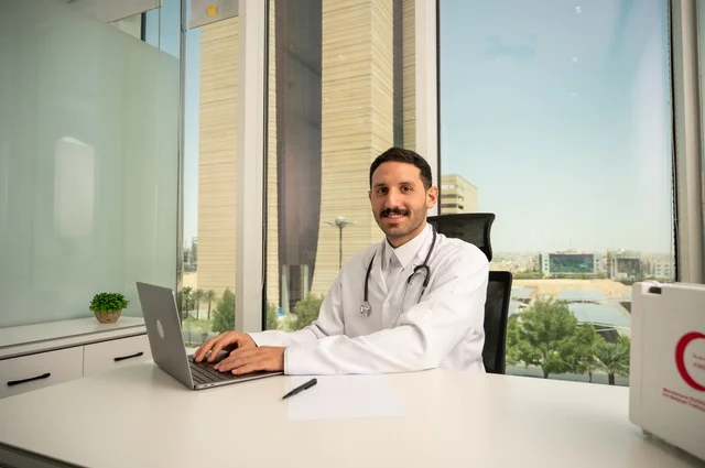 تسجيل التقارير والحالات الصحيه، متابعة حالات المرضى عبر الاجهزة و التقنيات الحديثة, استخدام حقيبة الاسعافات الاولية وجهاز الحاسوب في الطب في تسجيل الحالات، طبيب عربي خليجي سعودي مبتسم يرتدي المعطف الأبيض يجلس في عيادته وينظر إلى الكاميرا بإيماءات الثقة بالنفس