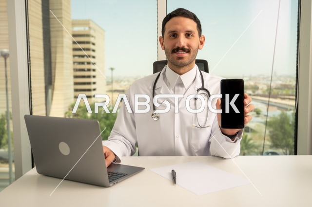 عرض شاشة سوداء فارغة عبر الهاتف الجوال، النظر إلى الكاميرا بإيماءات السرور، العمل في القطاعات الصحية، استخدام جهاز الحاسوب في الطب في تسجيل الحالات، تسجيل التقارير والحالات الصحيه, طبيب عربي خليجي سعودي مبتسم يرتدي المعطف الأبيض يجلس في عيادته