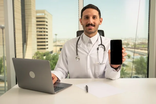 عرض شاشة سوداء فارغة عبر الهاتف الجوال، النظر إلى الكاميرا بإيماءات السرور، العمل في القطاعات الصحية، استخدام جهاز الحاسوب في الطب في تسجيل الحالات، تسجيل التقارير والحالات الصحيه, طبيب عربي خليجي سعودي مبتسم يرتدي المعطف الأبيض يجلس في عيادته