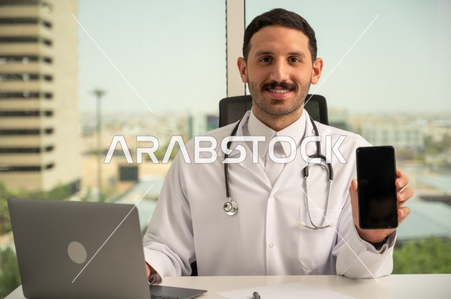 عرض شاشة سوداء فارغة عبر الهاتف الجوال، النظر إلى الكاميرا بإيماءات السرور، العمل في القطاعات الصحية، استخدام جهاز الحاسوب في الطب في تسجيل الحالات، تسجيل التقارير والحالات الصحيه, طبيب عربي خليجي سعودي مبتسم يرتدي المعطف الأبيض يجلس في عيادته