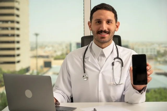 عرض شاشة سوداء فارغة عبر الهاتف الجوال، النظر إلى الكاميرا بإيماءات السرور، العمل في القطاعات الصحية، استخدام جهاز الحاسوب في الطب في تسجيل الحالات، تسجيل التقارير والحالات الصحيه, طبيب عربي خليجي سعودي مبتسم يرتدي المعطف الأبيض يجلس في عيادته