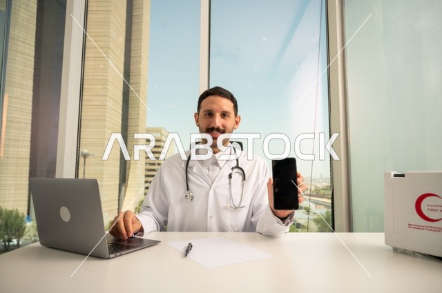 عرض شاشة سوداء فارغة عبر الهاتف الجوال، النظر إلى الكاميرا بإيماءات السرور، العمل في القطاعات الصحية، استخدام جهاز الحاسوب في الطب في تسجيل الحالات، تسجيل التقارير والحالات الصحيه, طبيب عربي خليجي سعودي مبتسم يرتدي المعطف الأبيض يجلس في عيادته