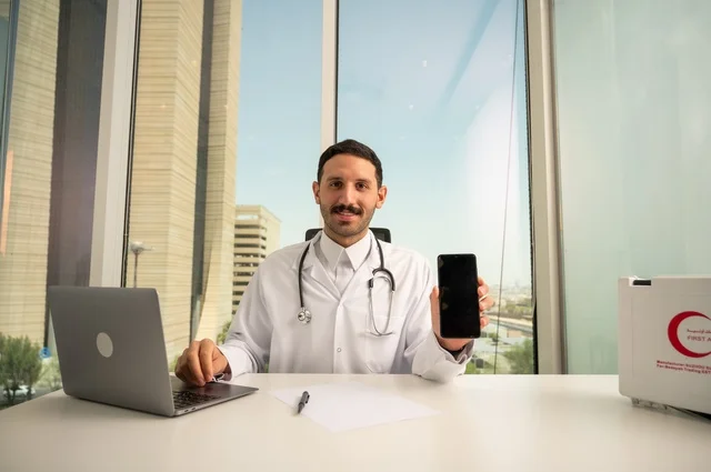 عرض شاشة سوداء فارغة عبر الهاتف الجوال، النظر إلى الكاميرا بإيماءات السرور، العمل في القطاعات الصحية، استخدام جهاز الحاسوب في الطب في تسجيل الحالات، تسجيل التقارير والحالات الصحيه, طبيب عربي خليجي سعودي مبتسم يرتدي المعطف الأبيض يجلس في عيادته