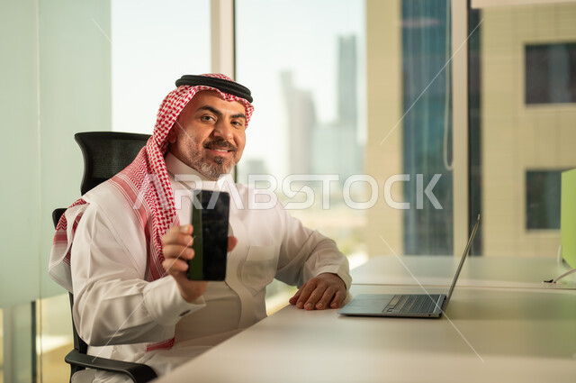 Blank black mobile phone screen, business management and organization, using modern and advanced technical device, office administrative profession and job, Saudi Arabian Gulf man wearing traditional thobe and shemagh working on laptop inside office, companies and offices in the Kingdom of Saudi Arabia