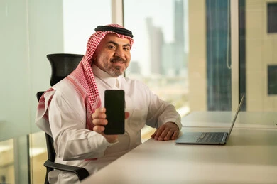 عرض شاشة هاتف محمول سوداء فارغة, إدارة شؤون الأعمال وتنظيمها، استخدام جهاز تقني حديث ومتطور، مهنة ووظيفة ادارية مكتبية، رجل عربي خليجي سعودي يرتدي الثوب التقليدي والشماغ يقوم بالعمل على الحاسوب المحمول داخل المكتب، الشركات والمكاتب في المملكة العربية السعودية